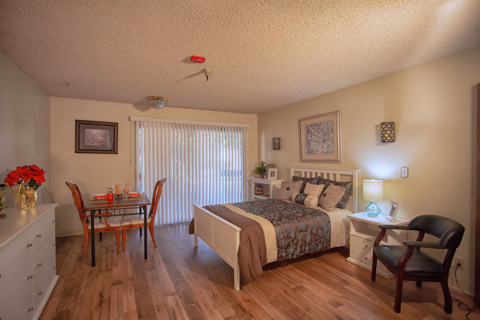 A cozy bedroom in a senior living facility featuring a white wooden bed with decorative pillows and a patterned bedspread. Next to the bed is a white nightstand with a lamp and framed photo. A black armchair is positioned nearby. On the left side of the room, there is a small dining table set for two with red chairs and tableware. A white dresser with a red flower arrangement sits against the wall. The room has wood flooring and a large sliding glass door with vertical blinds letting in natural light.