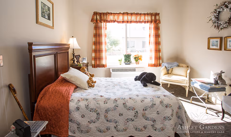 A cozy bedroom with a large wooden headboard bed covered in a patterned quilt and pillows, including teddy bears and a black stuffed animal. There is a window with plaid curtains letting in natural light, a small nightstand with a lamp and framed photos, a cane leaning against the wall, a cushioned chair, and a small table with books and a decorative rabbit. The room has light-colored walls and framed pictures hanging on them.