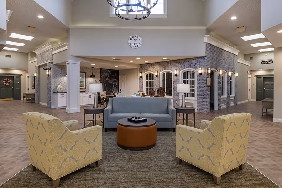 A spacious and well-lit common area in a senior living facility featuring a seating arrangement with a blue sofa, two yellow patterned armchairs, a round brown ottoman, and side tables with lamps. The room has a high ceiling with a large chandelier, a clock on the wall, and a brick wall with multiple windows and wall sconces. There are hallways leading to other rooms and a dining area visible in the background.