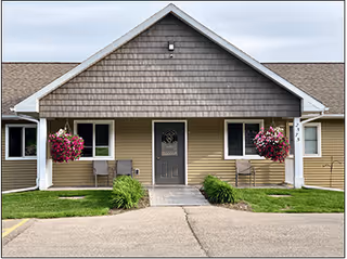 Front exterior view of a single-story building with a peaked roof, beige siding, and a central gray door flanked by two windows. Two hanging flower baskets with pink flowers are on either side of the entrance, and there is a paved walkway leading to the door. The building number 257 is visible on a white post to the right of the entrance.
