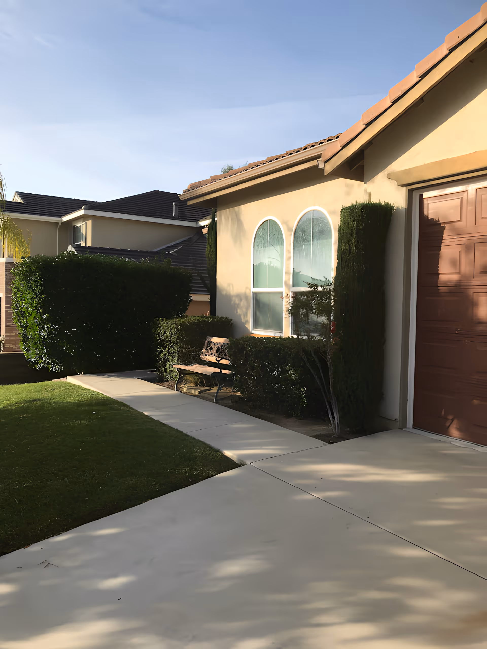 Front exterior of a single-story house showing a driveway and garage door, arched windows, trimmed hedges, and a bench by the walkway.
