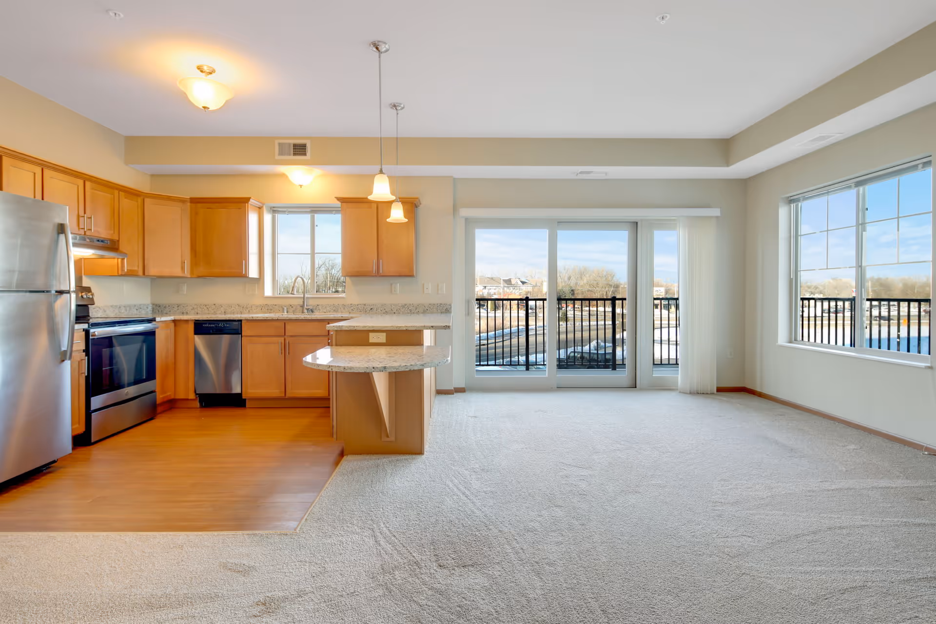 A bright and spacious open-concept kitchen and living area in a senior living facility. The kitchen features wooden cabinets, stainless steel appliances including a refrigerator, stove, and dishwasher, and granite countertops with a small breakfast bar. The living area has light-colored carpet and large windows with sliding glass doors that open to a balcony overlooking an outdoor view with trees and water.
