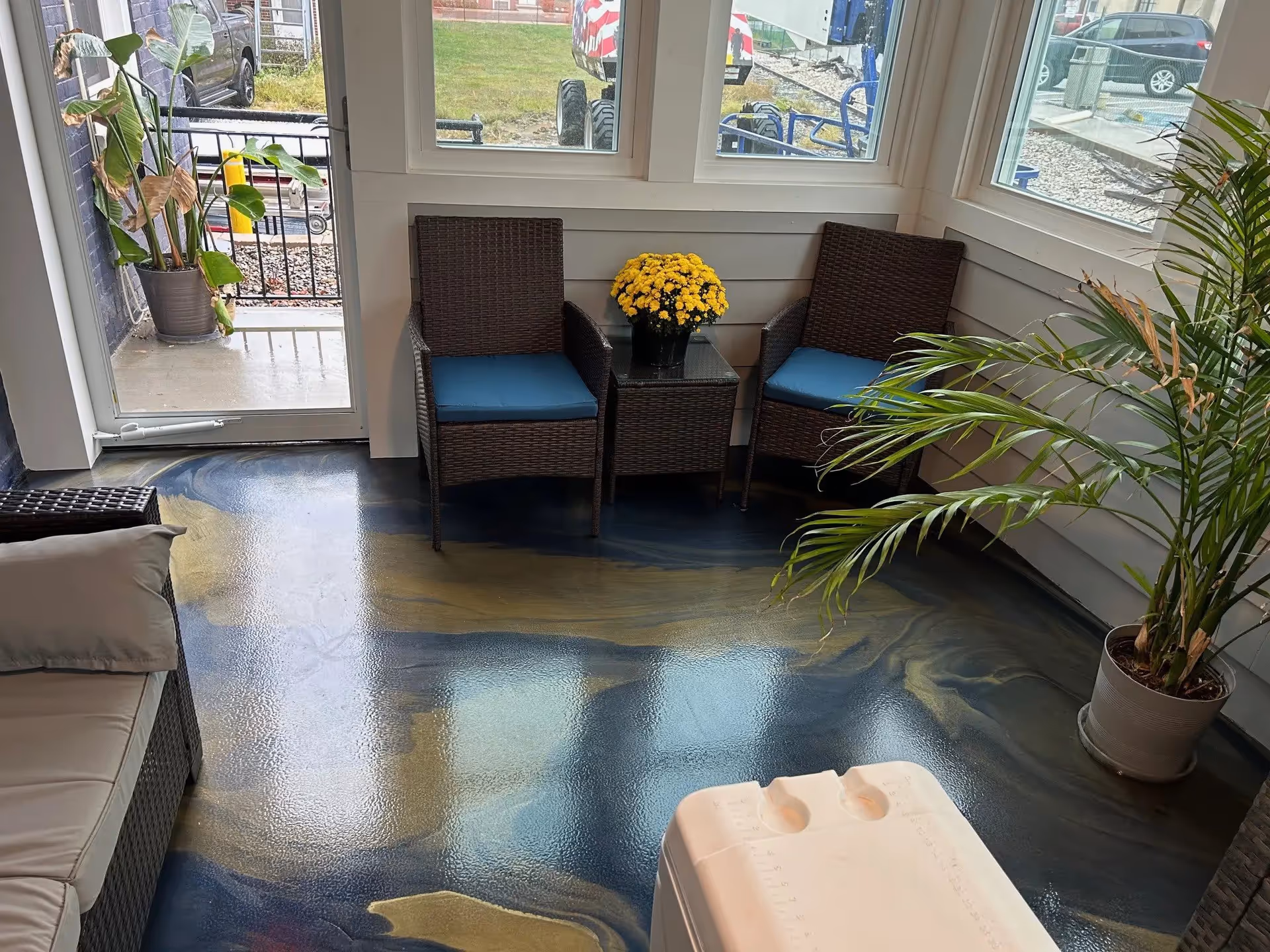 A cozy indoor seating area with two wicker chairs with blue cushions, a small glass-top table with a yellow flower pot, a beige cushioned sofa, and two potted plants. The floor has a glossy, marbled blue and gold finish. Large windows and a glass door provide a view of the outdoor area with grass and parked vehicles.