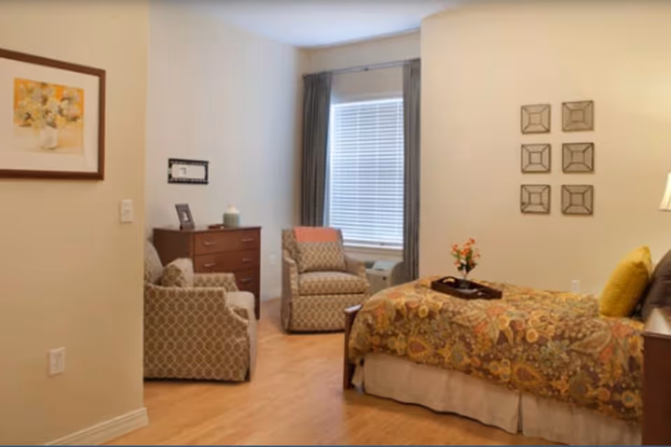 A cozy bedroom with a bed covered in a floral patterned bedspread, two cushioned armchairs with geometric patterns, a wooden dresser, a window with blinds and curtains, wall decorations including framed artwork and square mirrors, and a small table with a lamp and a flower vase.