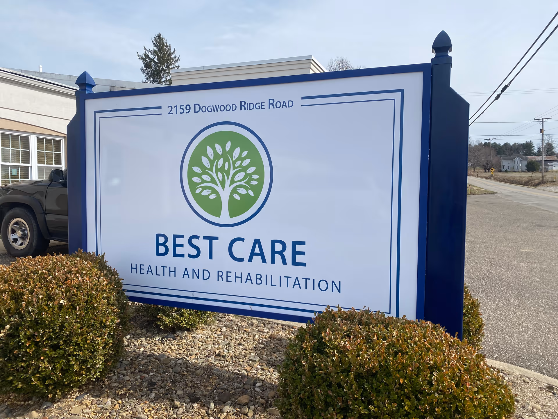 Outdoor sign with a green tree logo reading "BEST CARE Health and Rehabilitation" and the address 2159 Dogwood Ridge Road, set among shrubs by a driveway.