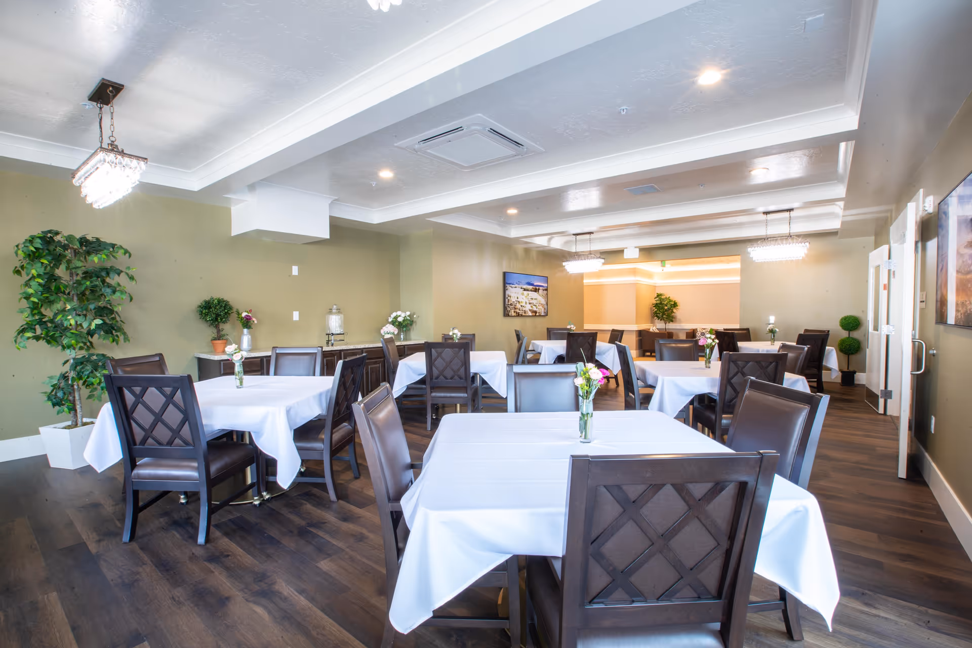 A bright and spacious dining room with multiple square tables covered with white tablecloths, each adorned with a small vase of flowers. The room features dark wooden chairs with cushioned seats, light green walls, wood flooring, and modern rectangular chandeliers hanging from a white ceiling. There are potted plants and framed artwork on the walls, creating a welcoming atmosphere.