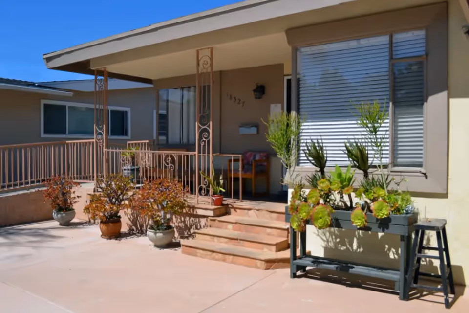 Front porch of a residential care facility with steps leading up to a covered entrance. Several potted plants and a planter with succulents are arranged around the porch. The building has beige walls and a large window with closed blinds.