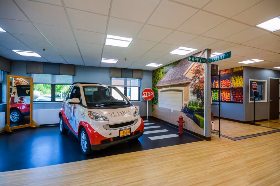 Indoor area designed to resemble a small street scene with a red and white compact car labeled 'St. James Rehabilitation & Healthcare Center' parked on a black floor with white pedestrian crossing lines. The walls feature murals of a house, a stop sign, a fire hydrant, and a fruit market. There are street signs labeled 'Happy Ave' and 'Healthy St' mounted on a pole. The room has large windows, wood flooring, and bright overhead lighting.