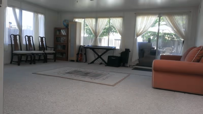 Spacious carpeted living room with an orange sofa, wooden chairs, area rug, keyboard and bookshelf near a sliding glass door with sheer curtains.