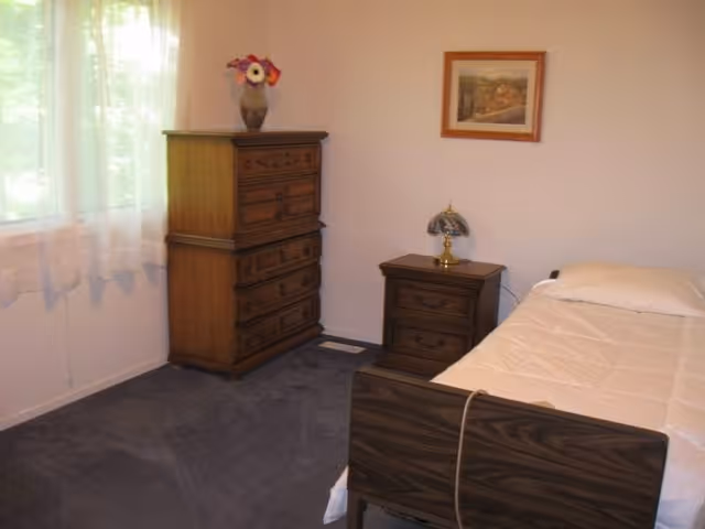 A small bedroom with a single bed covered with a white comforter, a wooden nightstand with a decorative lamp, a wooden dresser with a vase of flowers on top, a framed picture on the wall, and a window with sheer white curtains letting in natural light.