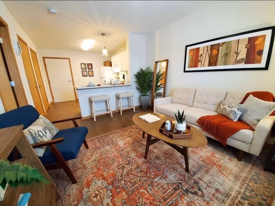 A cozy living room with a beige sofa adorned with pillows and an orange throw blanket, a wooden coffee table with decorative items, a blue armchair with a pillow, a large patterned rug, a tall mirror, and a potted plant. In the background, there is a kitchen area with white cabinets, a refrigerator, and a counter with two stools.