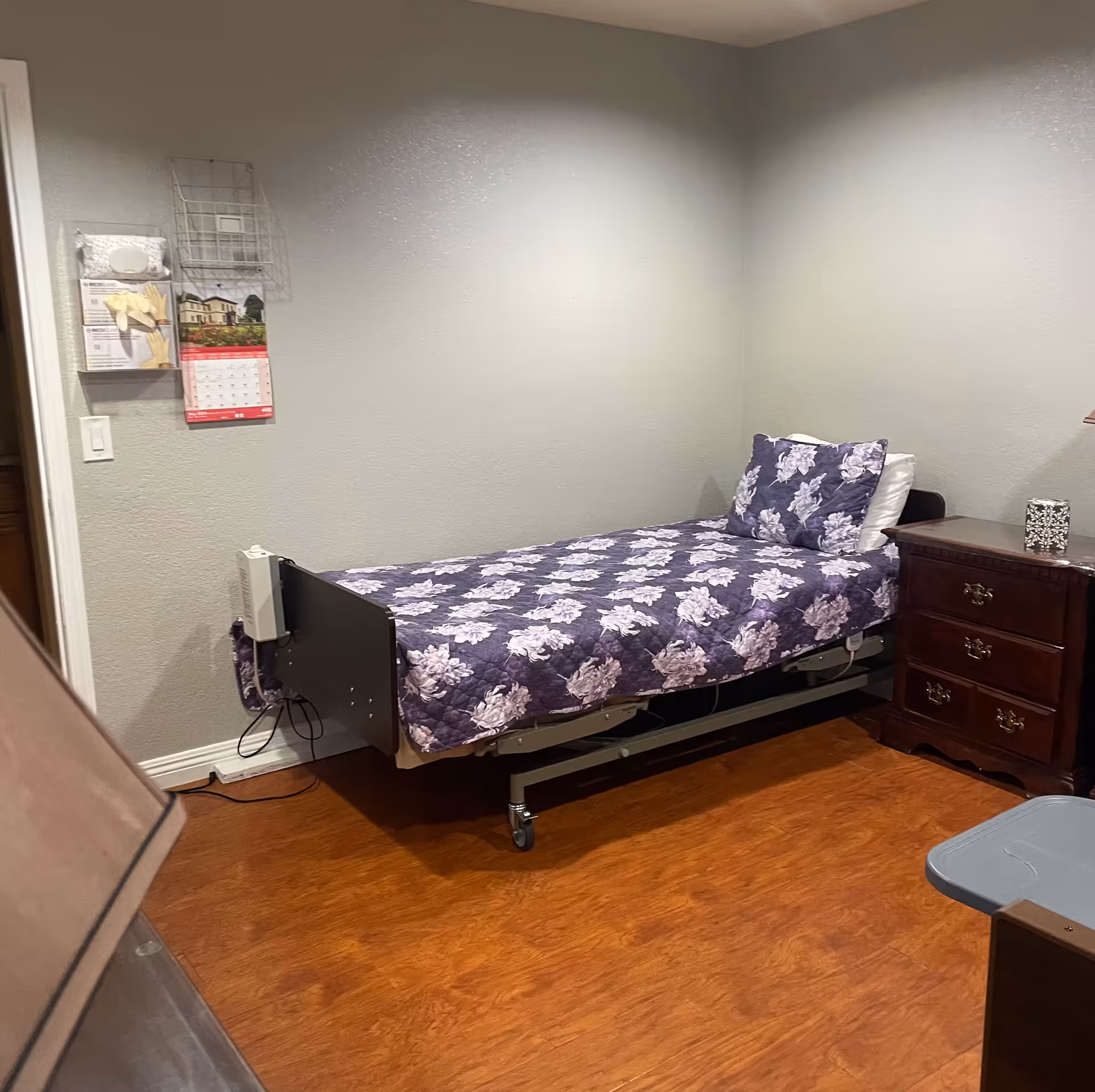 A small bedroom with a single bed covered in a purple floral quilt and matching pillow. Next to the bed is a wooden nightstand with three drawers and a decorative tissue box on top. The room has wooden flooring and light gray walls. On the left wall, there is a calendar and some medical supplies hanging. A lamp is partially visible in the foreground.