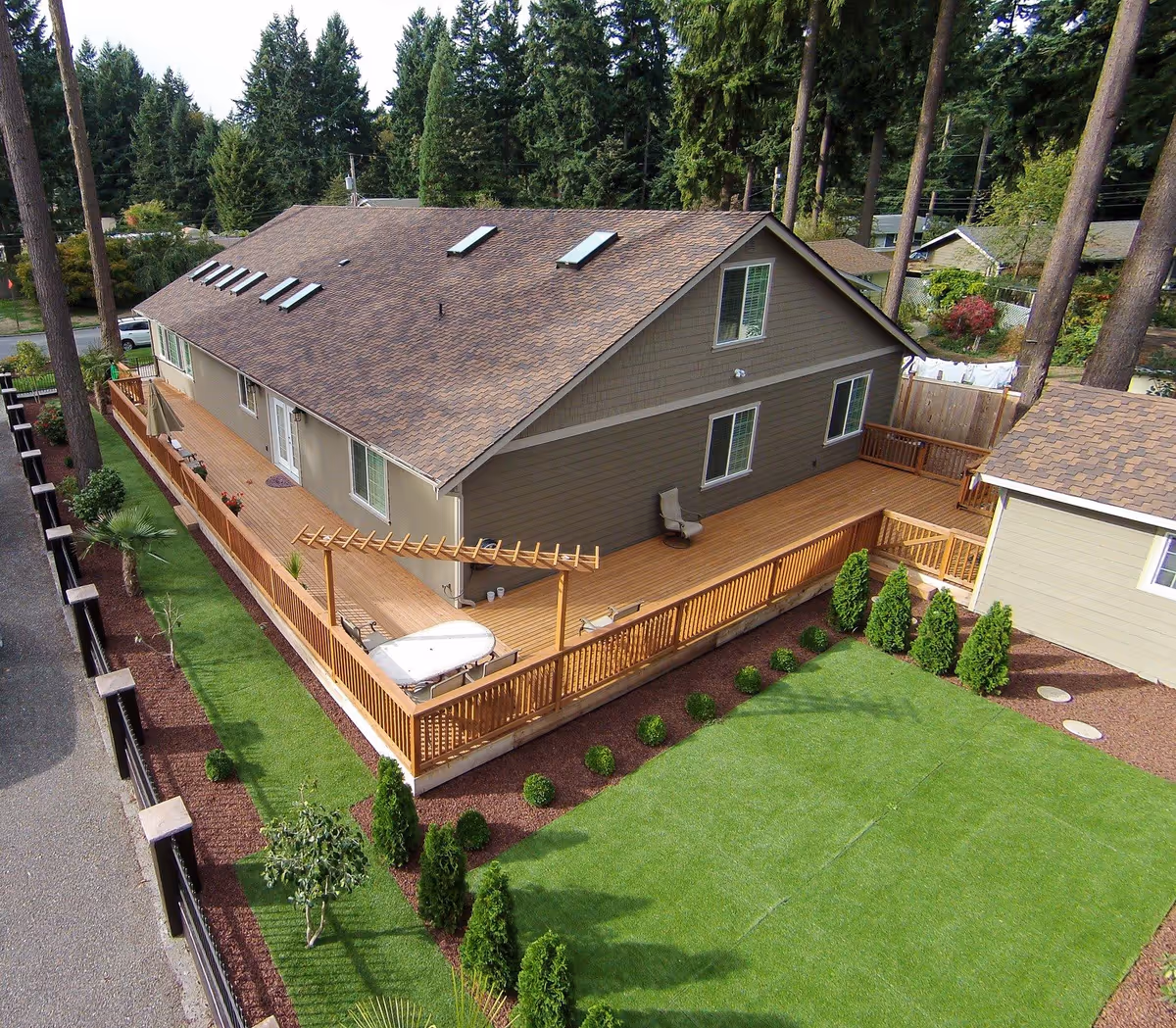 Aerial view of a single-story residential building with a large wooden deck surrounding it. The building has a brown shingled roof with multiple skylights and beige siding. The deck features a pergola, outdoor seating, and is bordered by a well-maintained lawn with small shrubs and trees. Tall pine trees and neighboring houses are visible in the background.