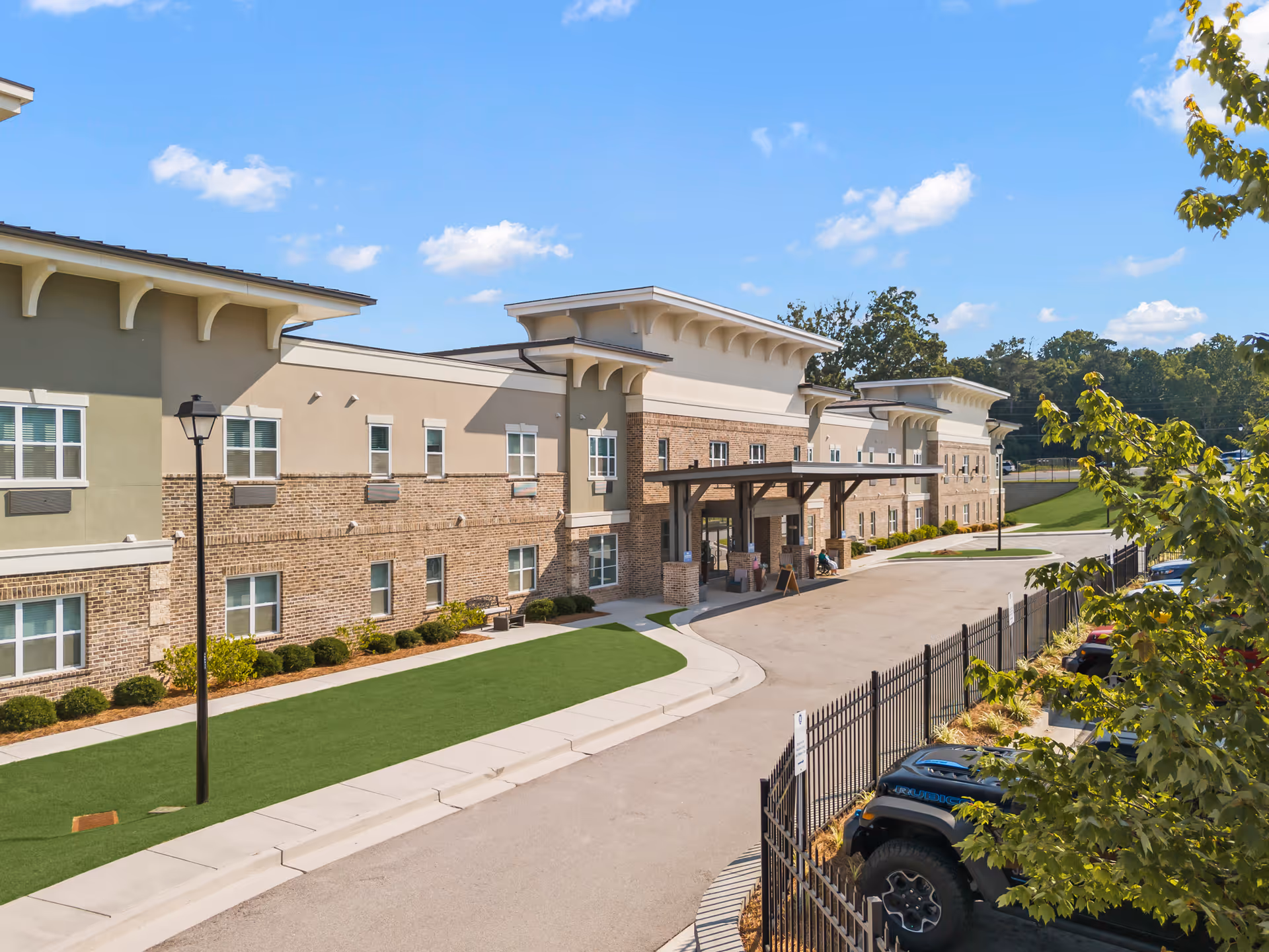 Front exterior of a two-story senior living facility with a covered entrance, landscaped lawn, driveway and parked cars.