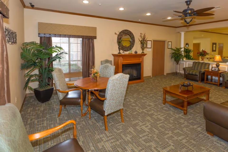 A cozy living room area in a senior living facility featuring a round wooden table with four upholstered chairs, a fireplace with decorative items on the mantel, a large potted plant near a window with curtains, a ceiling fan, and additional seating including armchairs and a coffee table on carpeted flooring.