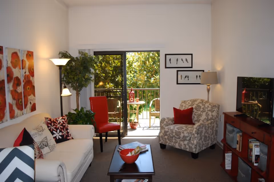 A cozy living room with a white sofa adorned with patterned pillows, a red chair, and a floral armchair with a red cushion. There is a coffee table with a red bowl and magazines, a floor lamp, and a TV on a red cabinet. The room has a sliding glass door leading to a balcony with outdoor seating and greenery visible outside. The walls are decorated with colorful floral artwork and framed bird pictures.