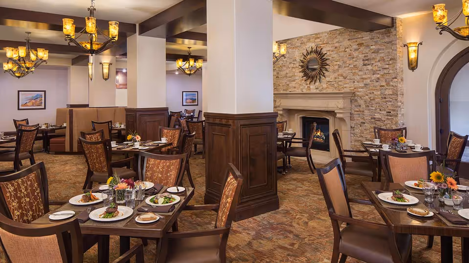 A warm and inviting dining room with multiple tables set for meals, featuring plates of food, glasses, and floral centerpieces. The room has elegant wooden chairs with patterned upholstery, a stone fireplace with a decorative sunburst mirror above it, and soft lighting from chandeliers and wall sconces.