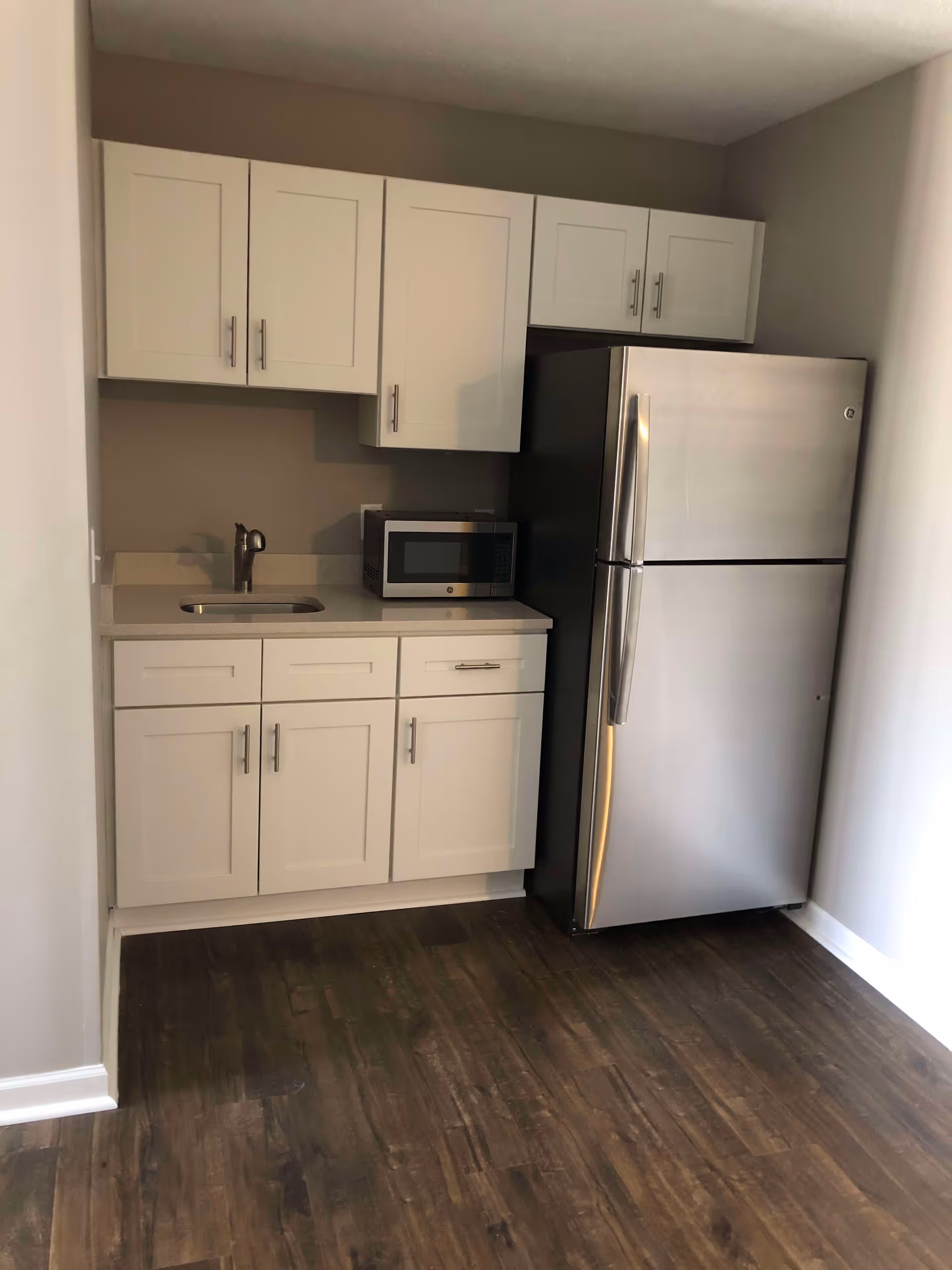 A small kitchen area with white cabinets, a stainless steel refrigerator, a microwave on the countertop, and a sink with a faucet. The floor is dark wood, and the walls are painted light gray.