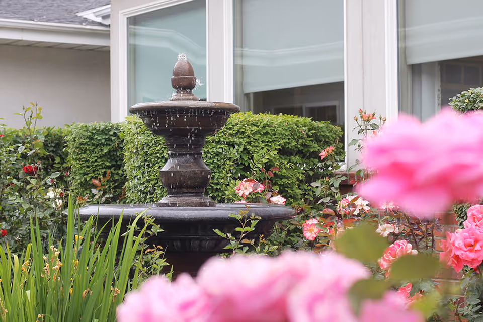 A tiered stone fountain surrounded by pink roses and green shrubs in front of a building with windows.