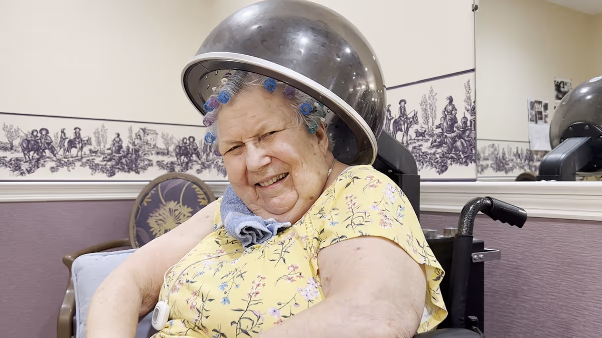 An elderly woman sitting in a wheelchair under a hair dryer in a salon area, smiling and wearing a yellow floral shirt with hair curlers in her hair. The background shows a chair, a wall with decorative wallpaper, and a large mirror reflecting part of the scene.