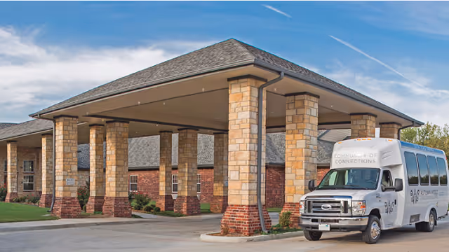 Entrance of a senior living facility with a covered drop-off area supported by stone pillars. A white shuttle bus labeled 'Community of Connections' is parked near the entrance. The building has brick and stone exterior walls and a gray shingled roof under a partly cloudy blue sky.
