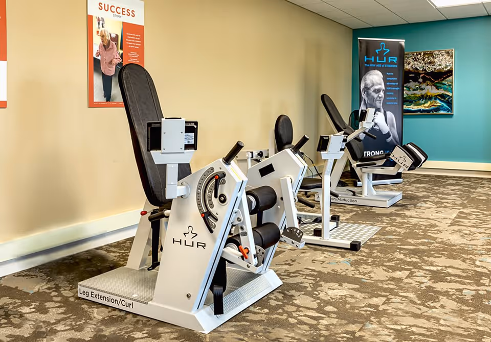 A fitness room with two HUR exercise machines designed for leg extension and curl, and hip abduction and adduction. The room has beige and teal walls, a patterned carpet, and motivational posters on the walls.