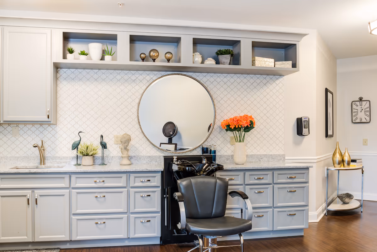 A salon area with a black styling chair in front of a round mirror mounted on a white tiled wall. Below the mirror is a black sink for washing hair. Surrounding the mirror are light gray cabinets and drawers with silver handles. On the countertop are decorative items including a vase with orange flowers, small sculptures, and plants. The floor is wooden, and there is a small table with gold vases and books in the background.