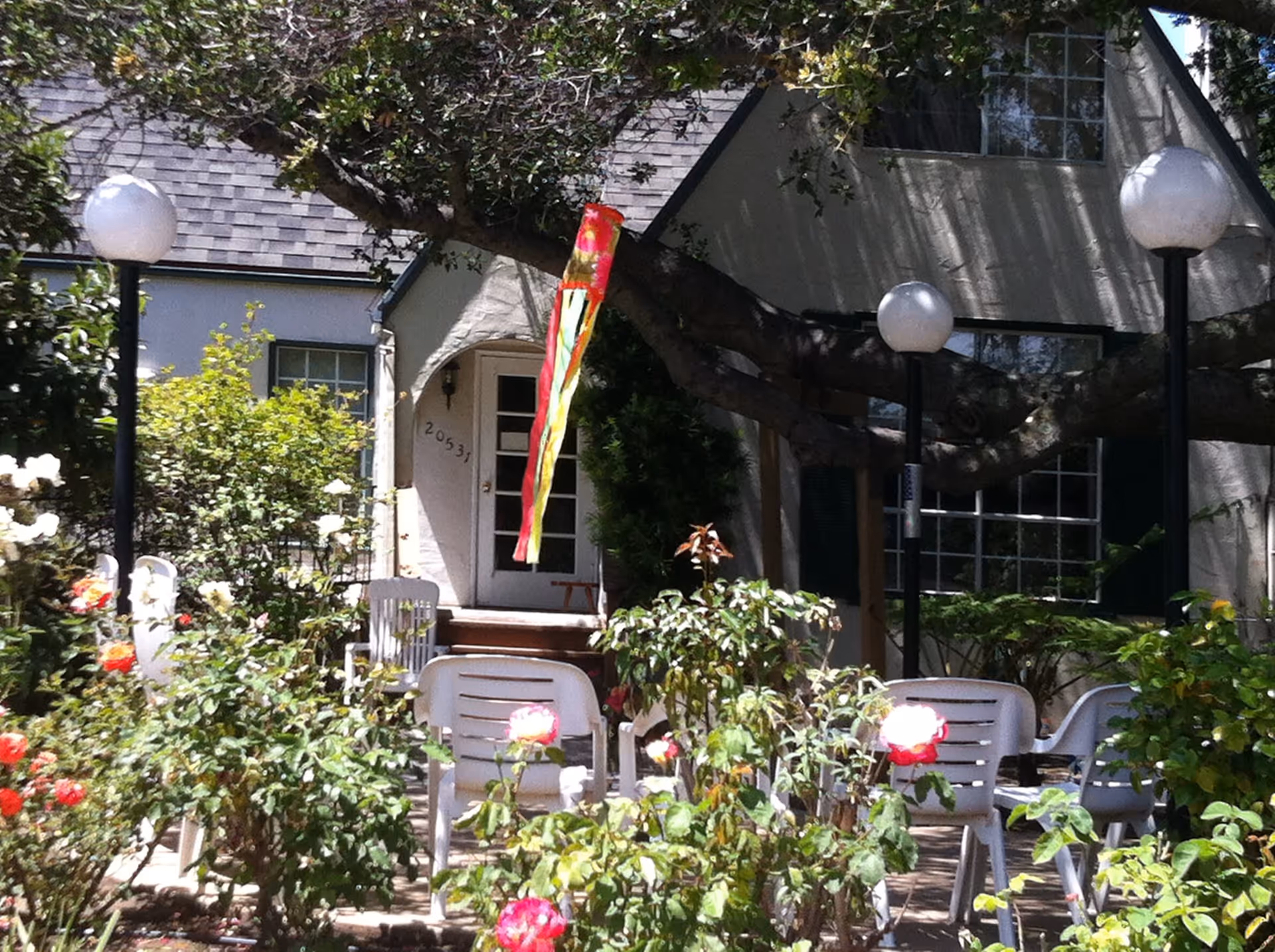 Front entrance of a cottage-style building with a garden of roses, plastic chairs, lamp posts and a colorful windsock hanging from a tree.