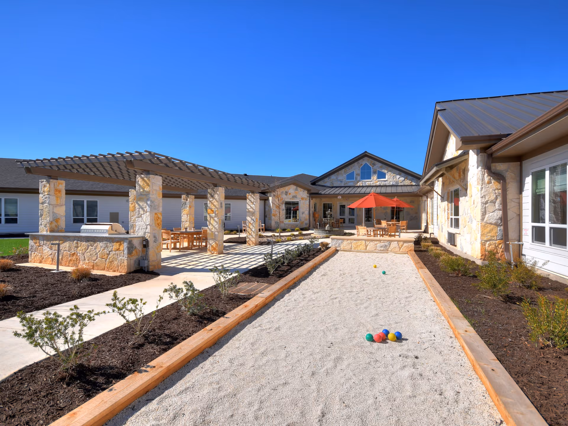 Outdoor courtyard area at Alexis Pointe of Wimberley Senior Living featuring a bocce ball court with colorful balls, stone pillars supporting a pergola with wooden tables and chairs underneath, a red umbrella shading a seating area, and a clear blue sky.