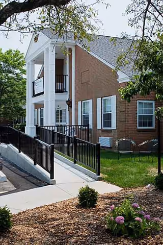 Brick senior living building with white columns, ramp, walkway, and landscaped lawn.