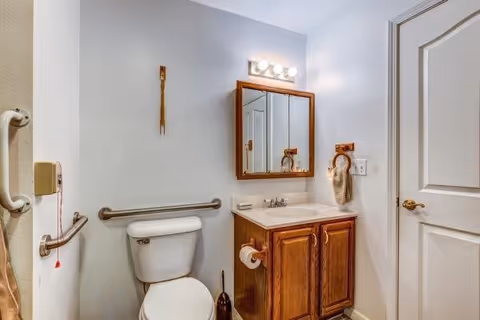 A small bathroom with a white toilet, wooden vanity with a sink, and a mirrored medicine cabinet above the sink. There are grab bars on the wall near the toilet and a towel ring with a beige towel next to the sink. The walls are painted light gray and there is a white door on the right side.