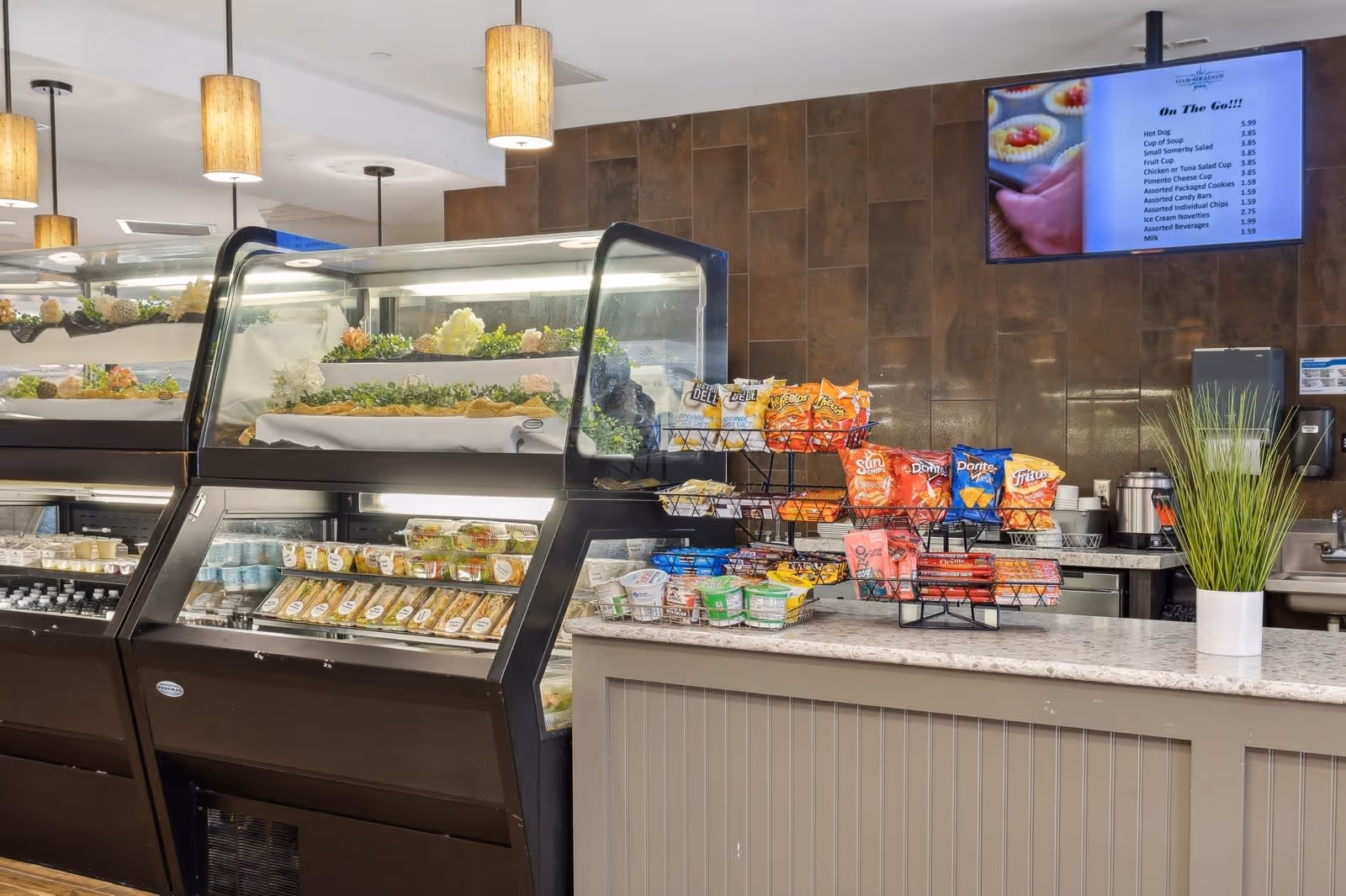 Interior view of a snack and salad bar area with refrigerated display cases containing pre-packaged salads and sandwiches. A countertop holds racks of assorted chips and snacks, with a potted plant on the right side. Pendant lights hang from the ceiling and a digital menu screen is mounted on the brown tiled wall in the background.