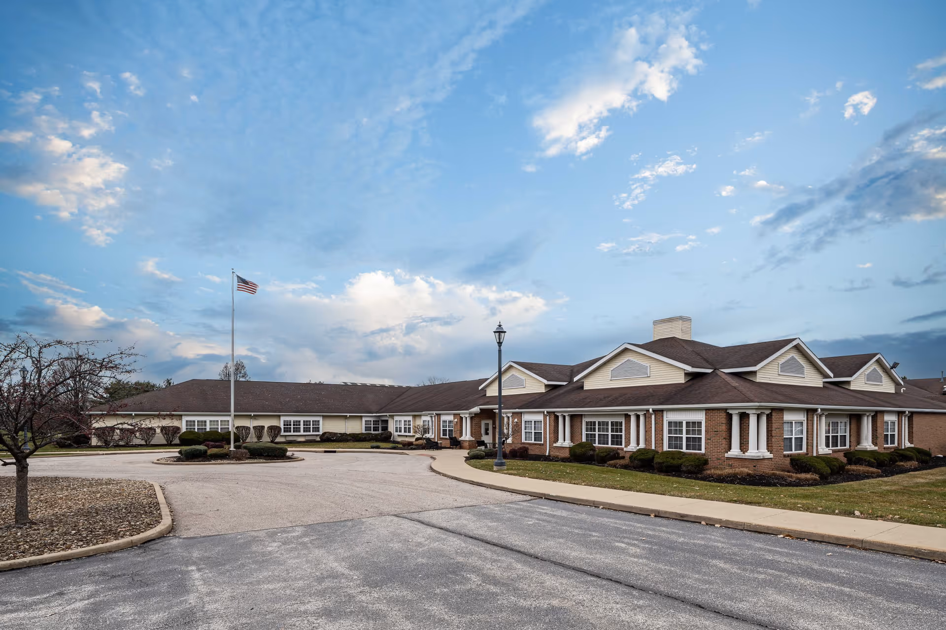 Front exterior of a single-story senior living building with a driveway, flagpole, and landscaped grounds under a blue sky.