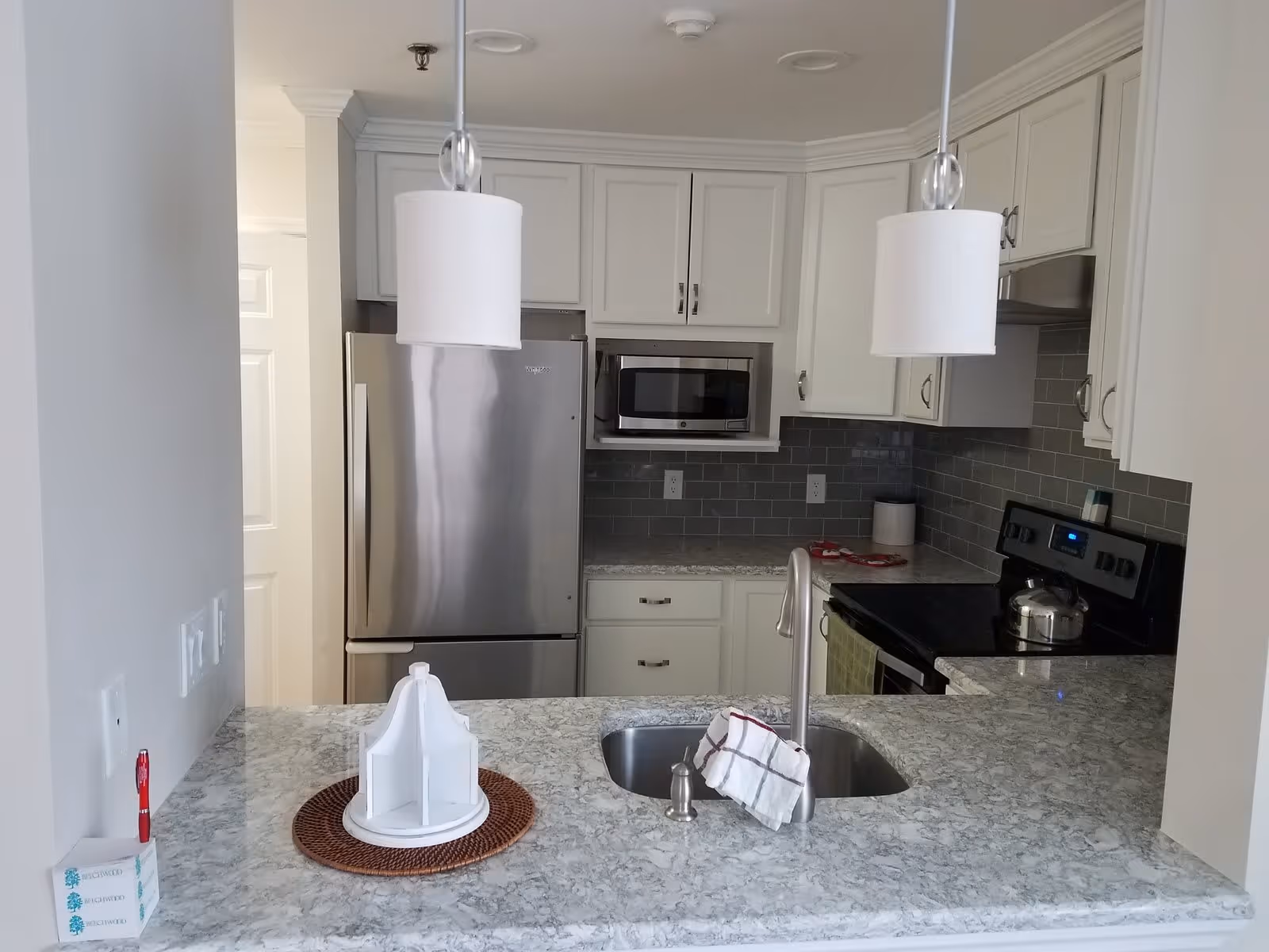 Modern kitchen with white cabinets, gray subway tile backsplash, stainless steel refrigerator and microwave, black stove with a kettle, and a marble countertop with a built-in sink and two hanging pendant lights above.