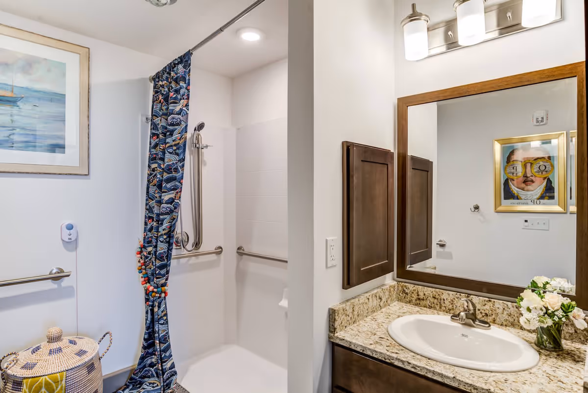 A bathroom with a walk-in shower featuring a blue patterned shower curtain and grab bars. There is a granite countertop with a white sink, a large mirror with a wooden frame, and a light fixture above it. A framed painting is visible on the wall, and a woven basket is placed near the shower.