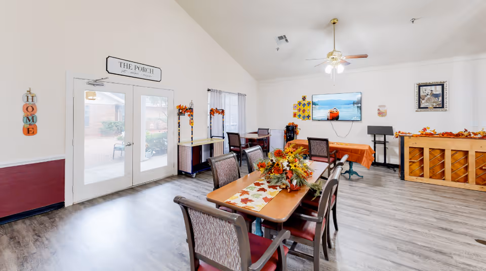 Bright communal dining and activity room with tables and chairs, fall decorations, a wall-mounted TV and double doors labeled "The Porch".