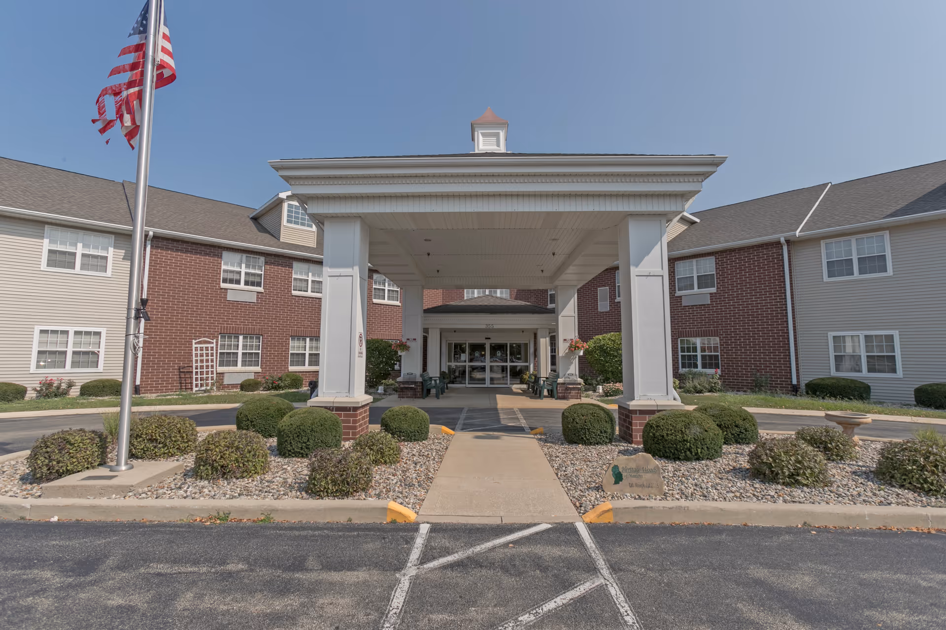 Front entrance of a senior living facility with a covered porte-cochere, American flag, and landscaped shrubs.