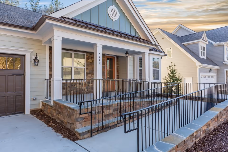 Front exterior view of a residential-style building with a covered porch, stone accents, a wooden door, and a wheelchair-accessible ramp with black metal railings leading up to the entrance.