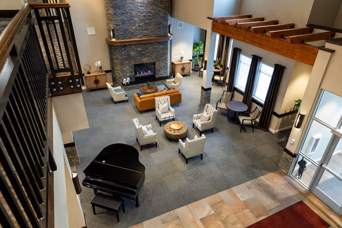 View from above of a spacious living room area in a senior living facility featuring a stone fireplace, a brown leather sofa, several upholstered armchairs arranged around a round ottoman, a black grand piano, and large windows with dark curtains letting in natural light.