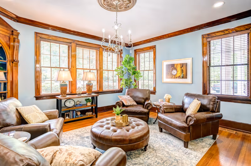 A cozy living room with light blue walls and wooden trim around multiple windows. The room features four brown leather armchairs arranged around a large round tufted leather ottoman on a patterned area rug. There is a wooden console table with two lamps and decorative items beneath the windows, a green leafy plant in the corner, and a framed picture on the wall. A chandelier hangs from the ceiling.