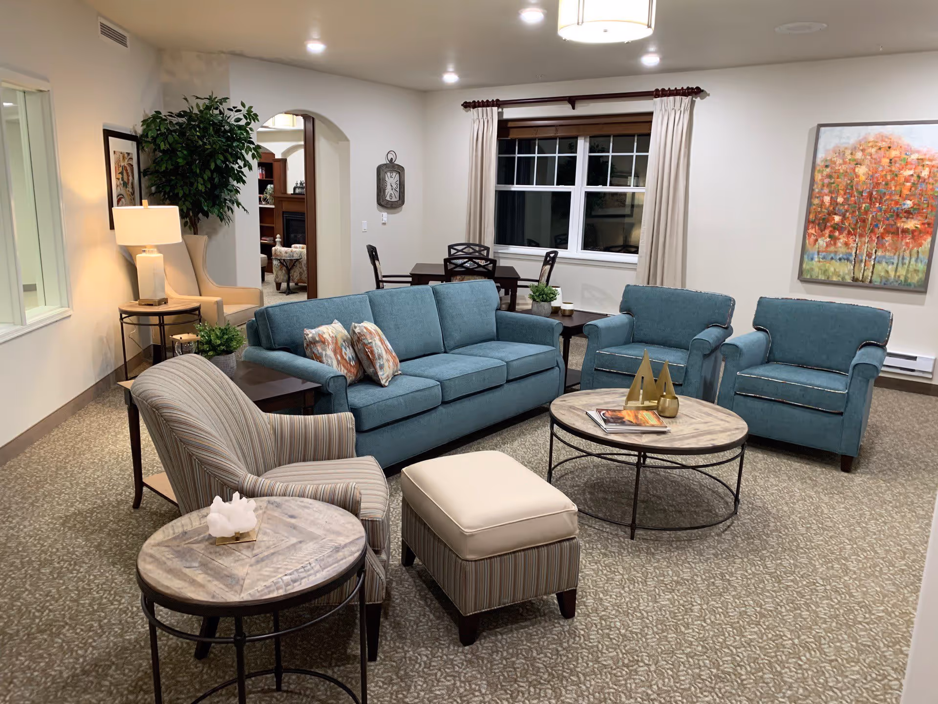 A well-lit communal living room with a teal sofa and matching armchairs arranged around round coffee tables and accent chairs.