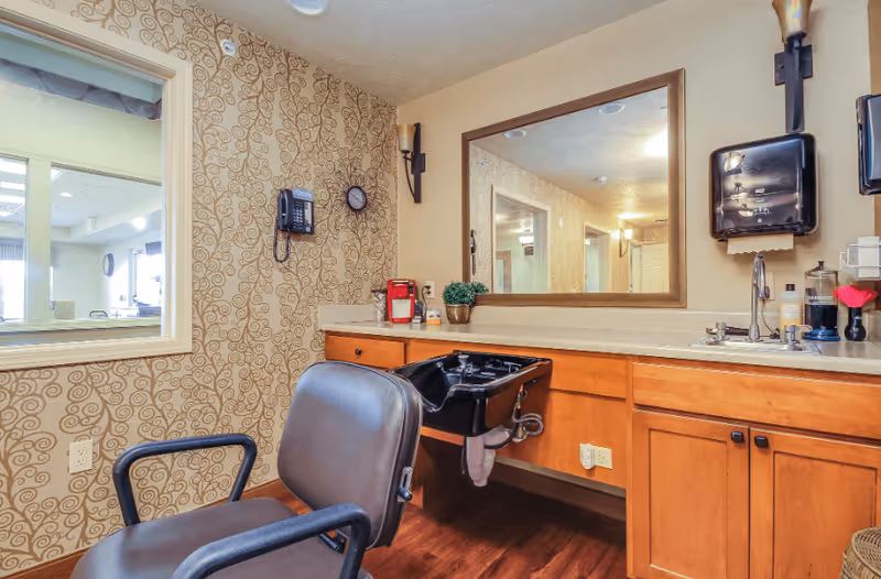 Interior view of a hair salon area in an assisted living facility featuring a brown salon chair in front of a black hair washing sink. The wall has patterned wallpaper with a mounted phone and clock. A large mirror is above a wooden counter with cabinets, a sink, and various hair care products. The floor is wooden and there is a window looking into another room.