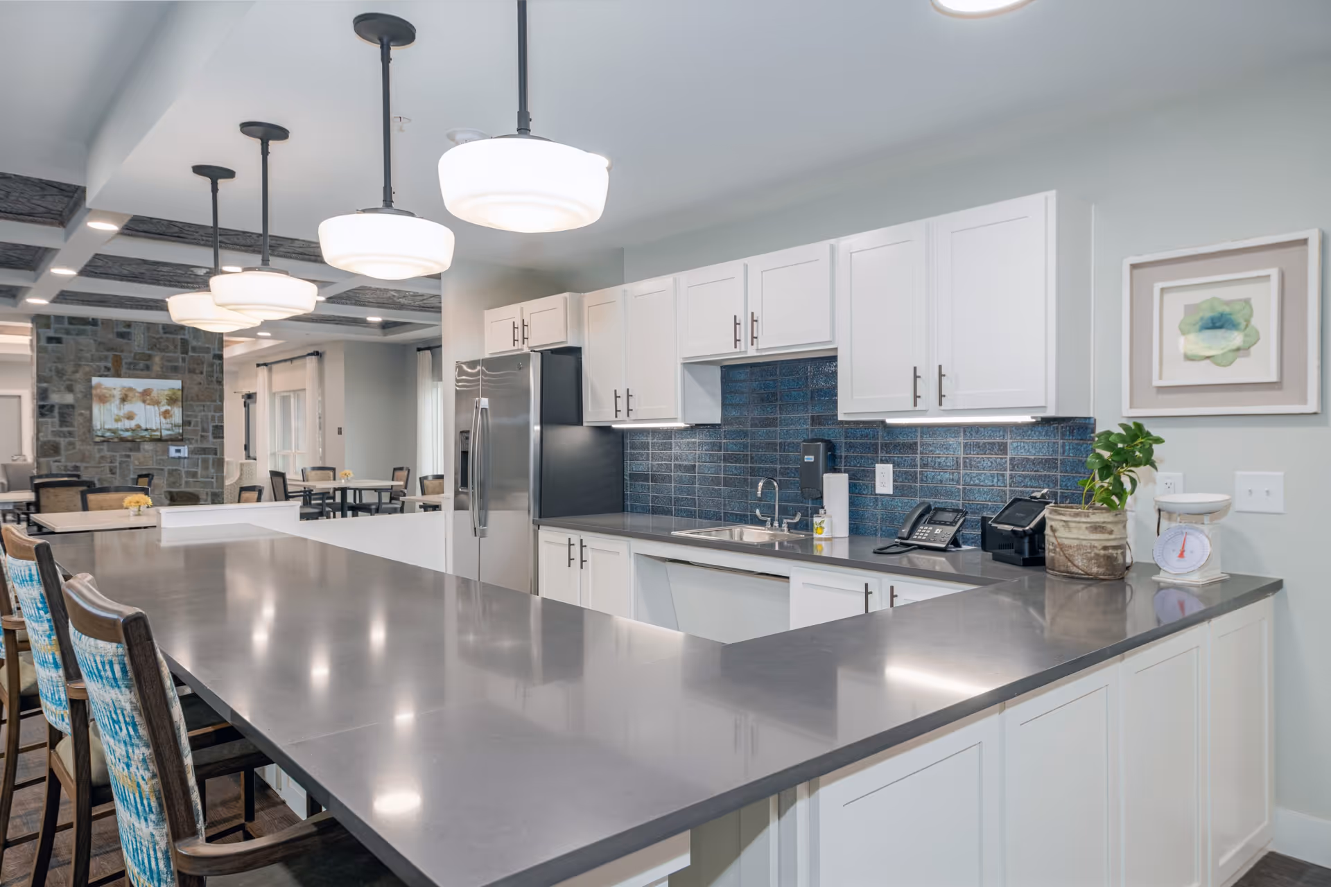 Modern kitchen area in a senior living facility with a long gray countertop, white cabinets, stainless steel refrigerator, blue tiled backsplash, and pendant lights hanging from the ceiling. There are chairs with patterned cushions along the counter and a dining area visible in the background.