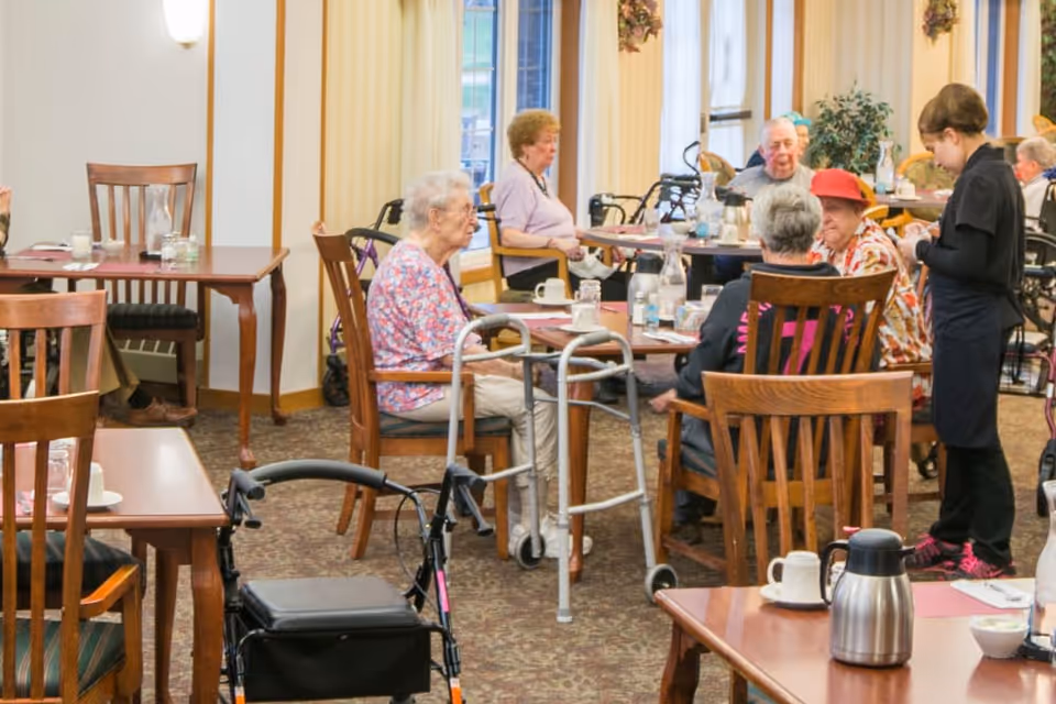 A dining room in a senior living facility with elderly residents seated at wooden tables. Some residents use walkers and wheelchairs. A staff member is attending to one of the tables. The room has large windows with vertical blinds and a carpeted floor.