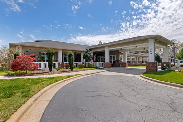 Driveway leading to a covered entrance and porch of a single-story senior living building with columns, landscaping, and a blue sky.