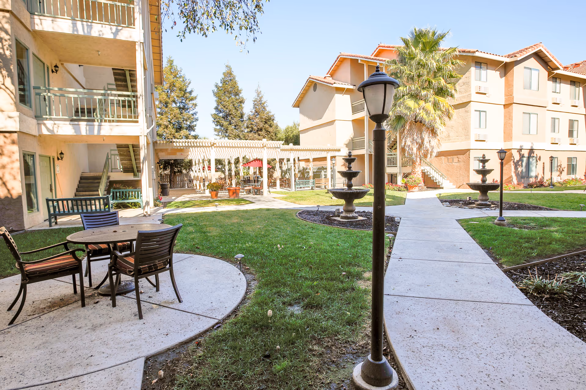 Outdoor courtyard area of a senior living facility with a round table and four chairs on a concrete patio. There are two multi-story buildings with balconies and staircases surrounding the grassy area. The courtyard features two tiered fountains, lamp posts, potted plants, and a pergola with seating underneath. Trees and clear blue sky are visible in the background.
