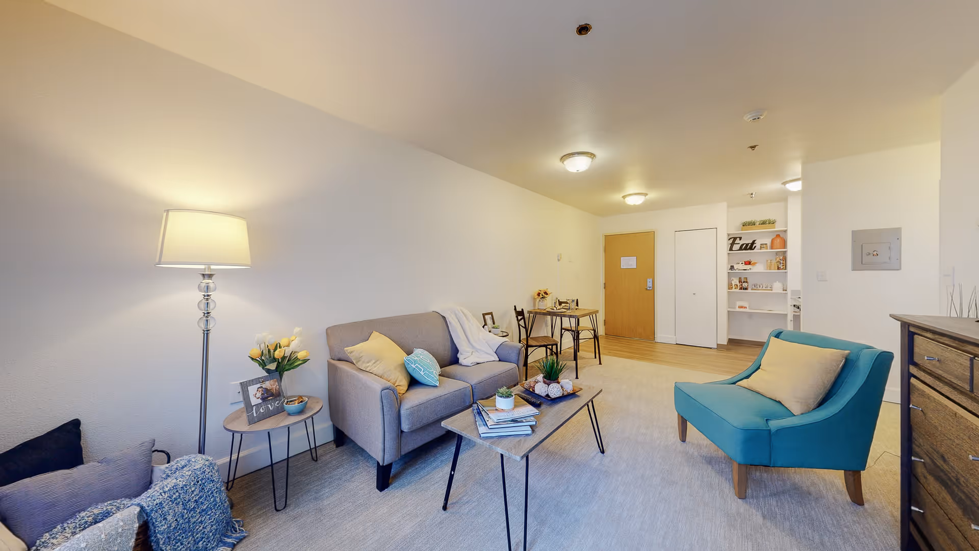 A bright and cozy living room in a senior living facility with a gray sofa adorned with cushions and a throw blanket, a teal armchair with a beige pillow, a wooden coffee table with books and small plants, a side table with a lamp, flowers, and a framed photo. In the background, there is a small dining table with two chairs, a wooden door, a white closet door, and built-in shelves with decorative items and the word 'Eat'.