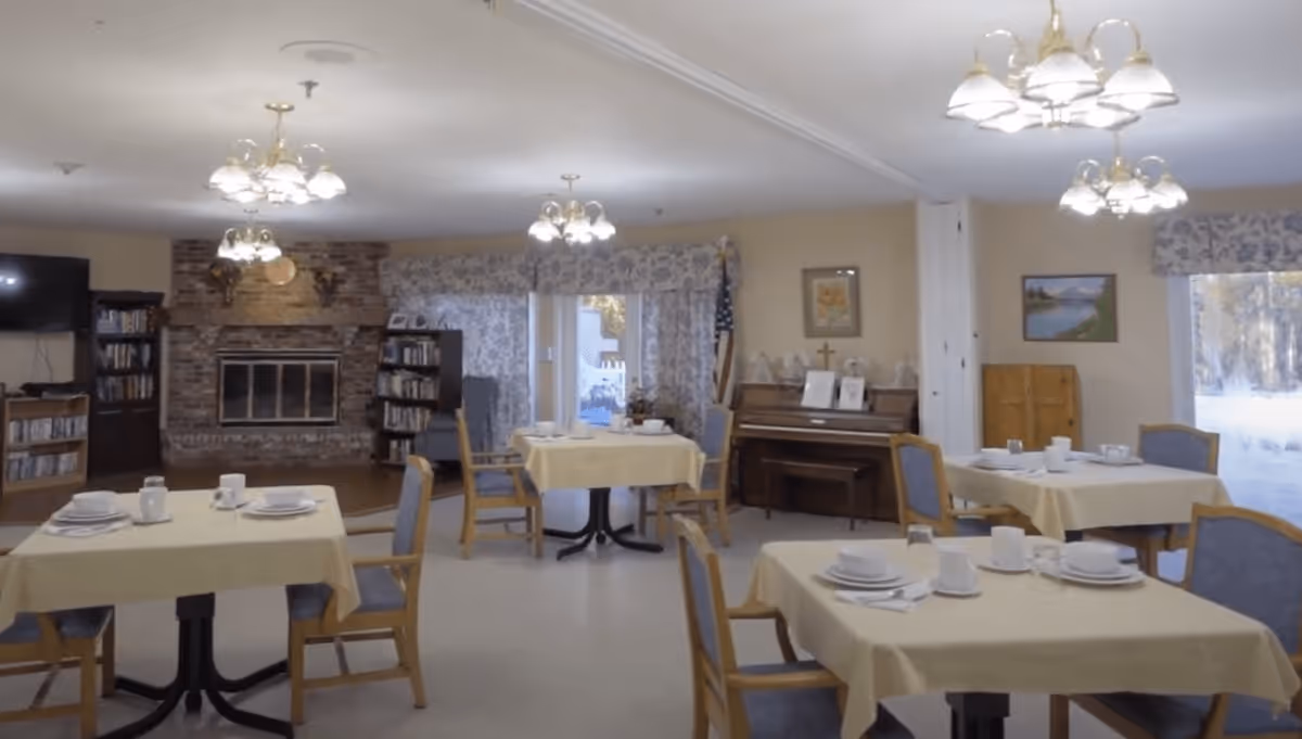 A dining room in the Norway Center facility with several tables covered with yellow tablecloths, each set with white plates, cups, and utensils. The room features wooden chairs with blue cushions, a brick fireplace, bookshelves, a piano, and large windows with floral curtains letting in natural light.