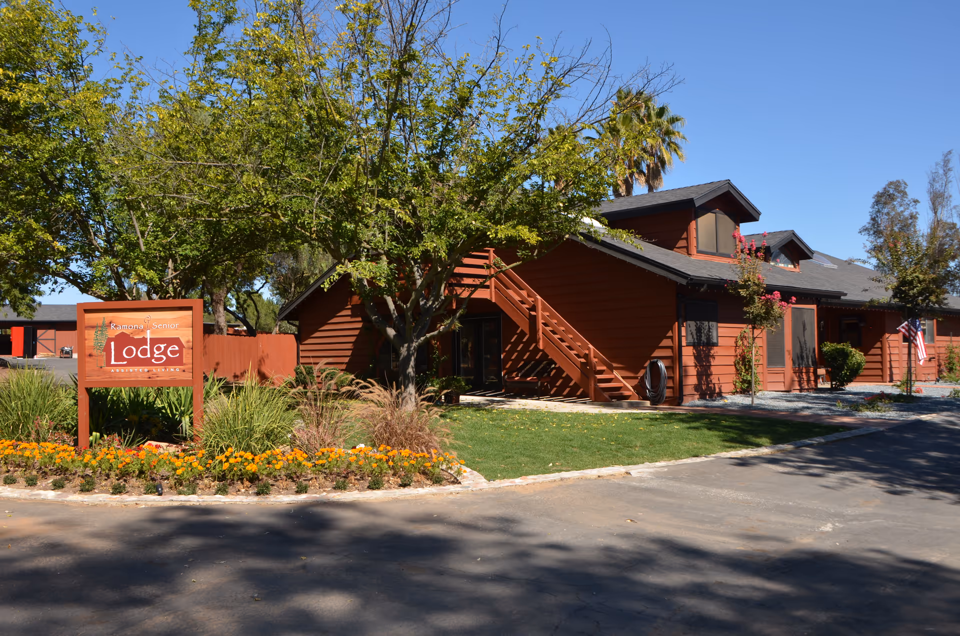 Exterior view of Ramona Senior Lodge, a single-story brown wooden building with a staircase leading to an upper level. The building is surrounded by green grass, trees, and a flower bed with orange flowers. A wooden sign in front reads 'Ramona Senior Lodge Assisted Living'. The sky is clear and blue.