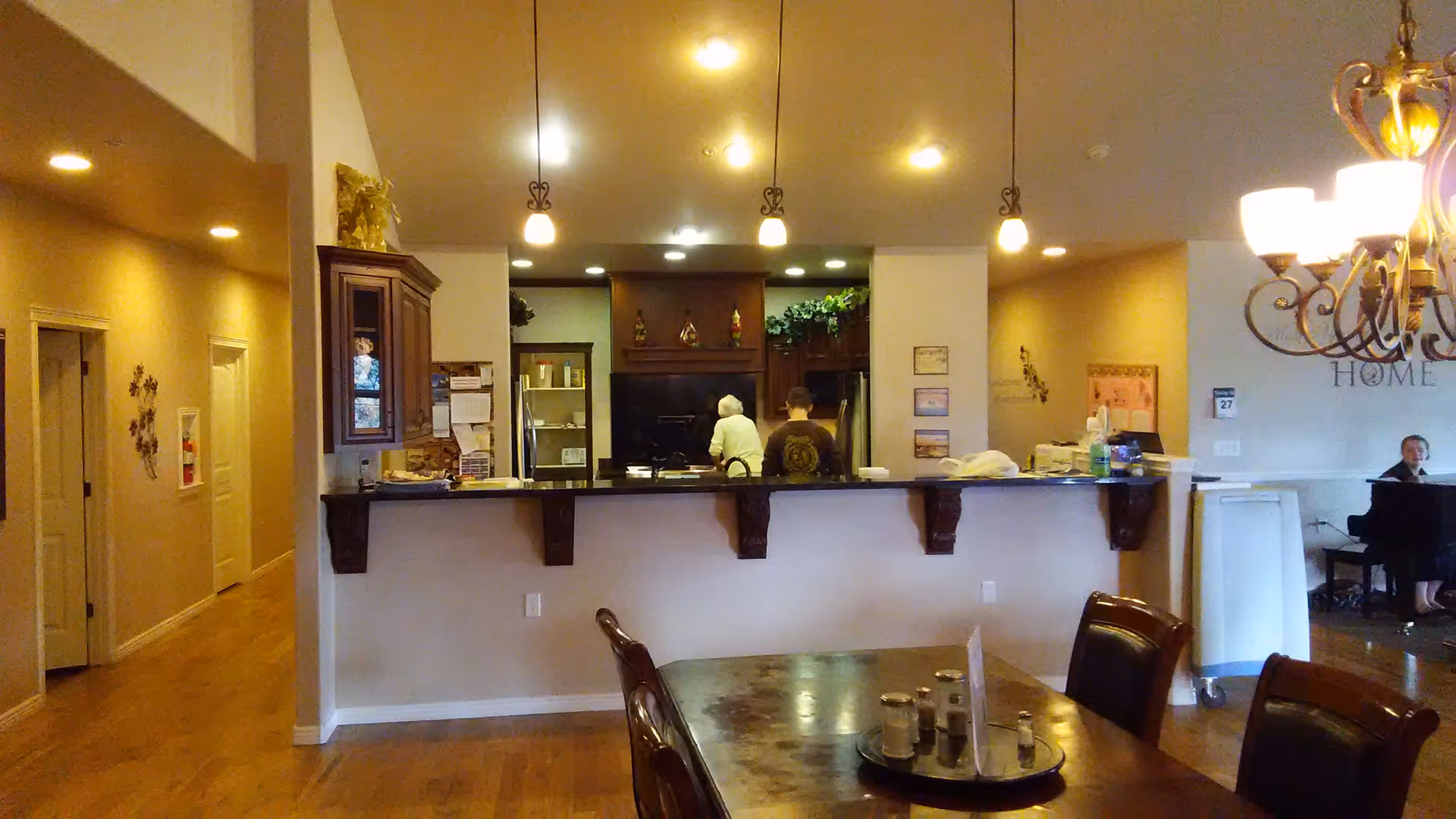 Interior view of a senior living facility showing a kitchen area with two people standing near the stove. In the foreground, there is a dining table with chairs and a chandelier hanging above. To the right, a person is seated at a piano. The space has warm lighting and wooden flooring.